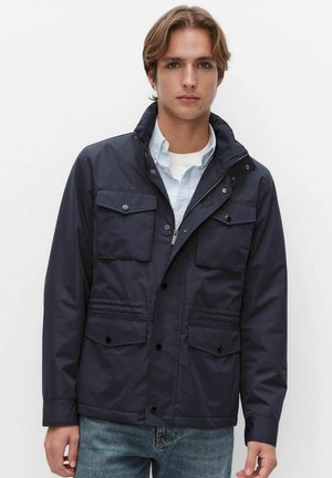 Jonge man met een marineblauw jack met vier voorzakken, een lichtblauw overhemd met knopen en een blauwe spijkerbroek, tegen een effen witte achtergrond.