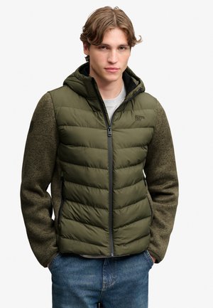 Junger Mann mit hellbraunen Haaren, der eine olivgrüne, gesteppte Jacke mit gestrickten Ärmeln und blaue Jeans trägt, Hände in den Taschen, nach vorne blickend.