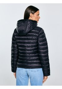 Sieviete ar tumšiem matiem, valkājot melnu jostoto kapuci ar kapuci un gaiši zilas džinsas, skats no sāniem pret vienkrāsainu fonu.