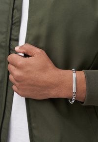Das silberne Armband verfügt über einen strukturierten rechteckigen Anhänger und runde Perlen, das am Handgelenk getragen wird. Im Hintergrund sind eine grüne Jacke und ein weißes Hemd zu sehen.
