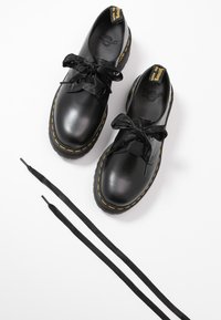 Paire de chaussures en cuir noir avec des semelles épaisses et des coutures jaunes, lacées avec des rubans noirs, avec des lacets plats noirs supplémentaires à côté.