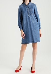 Denimová šaty s límečkem, dlouhými rukávy, zapínáním na knoflíky a kapsou na prsou. Mají volnější střih a modrou barvu. Červené boty doplňují vzhled.