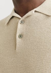 Camisa de polo de punto beige con un patrón texturizado, con un cuello y dos botones grises en la tapeta, vista en primer plano de la sección superior.