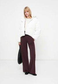 Witte pufferjas met capuchon, cropped model; crèmekleurige trui eronder, gecombineerd met donkerbruine wijd uitlopende broek en zwarte platte schoenen, draagt een zwarte tas.