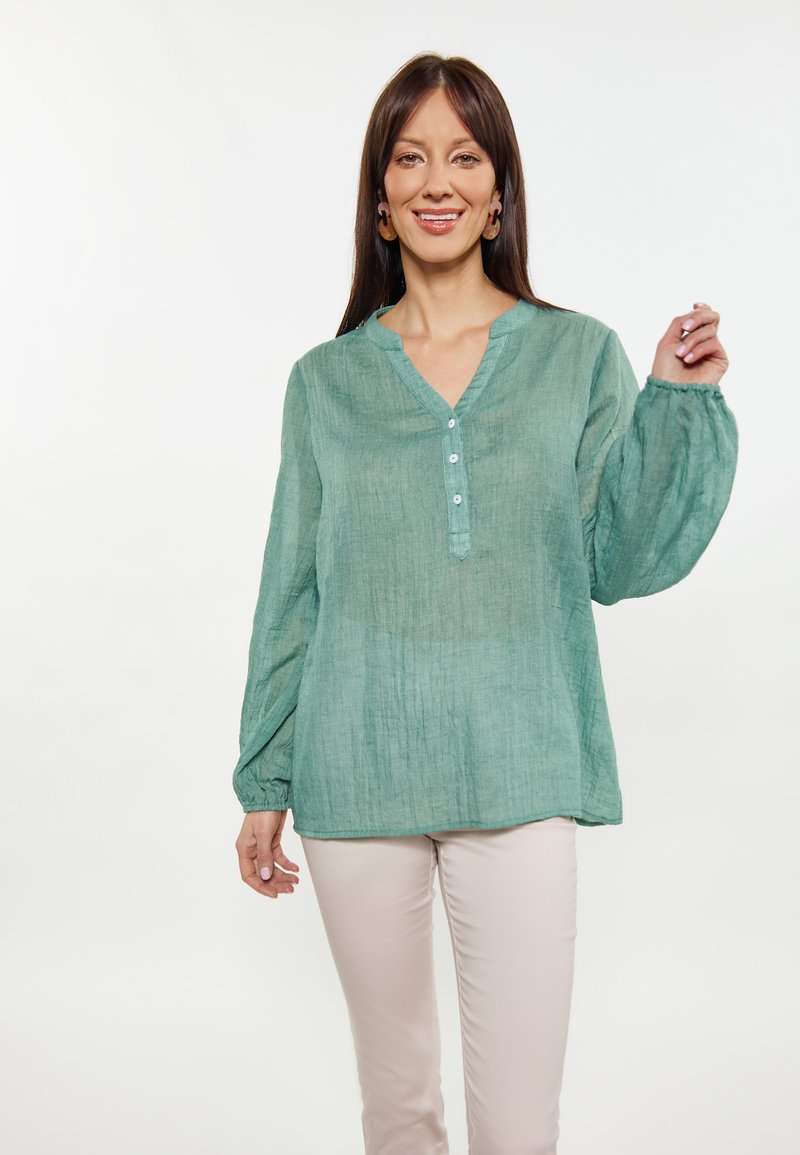 Blusa in lino verde chiaro con vestibilità comoda, maniche a sbuffo lunghe e patta con bottoni. Indossata con pantaloni chiari.