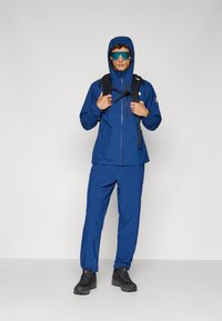 Μπλε αδιάβροχο μπουφάν με κουκούλα και φερμουάρ τσέπες, σε συνδυασμό με αντίστοιχα παντελόνια. Περιλαμβάνει μια μαύρη τσάντα πλάτης και γυαλιά προστασίας.