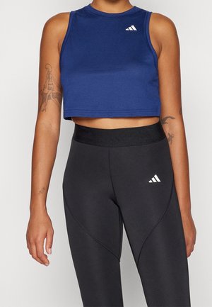 Osoba nosząca niebieski krótki top bez rękawów oraz czarne legginsy sportowe z wysokim stanem, oba z białymi logo Adidas, prezentująca tatuaże na ramionach.