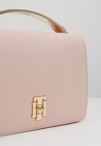 Sac à main en cuir rose blush avec poignée en métal doré et logo entrelacé "TH" en or sur le devant, sur un fond neutre.