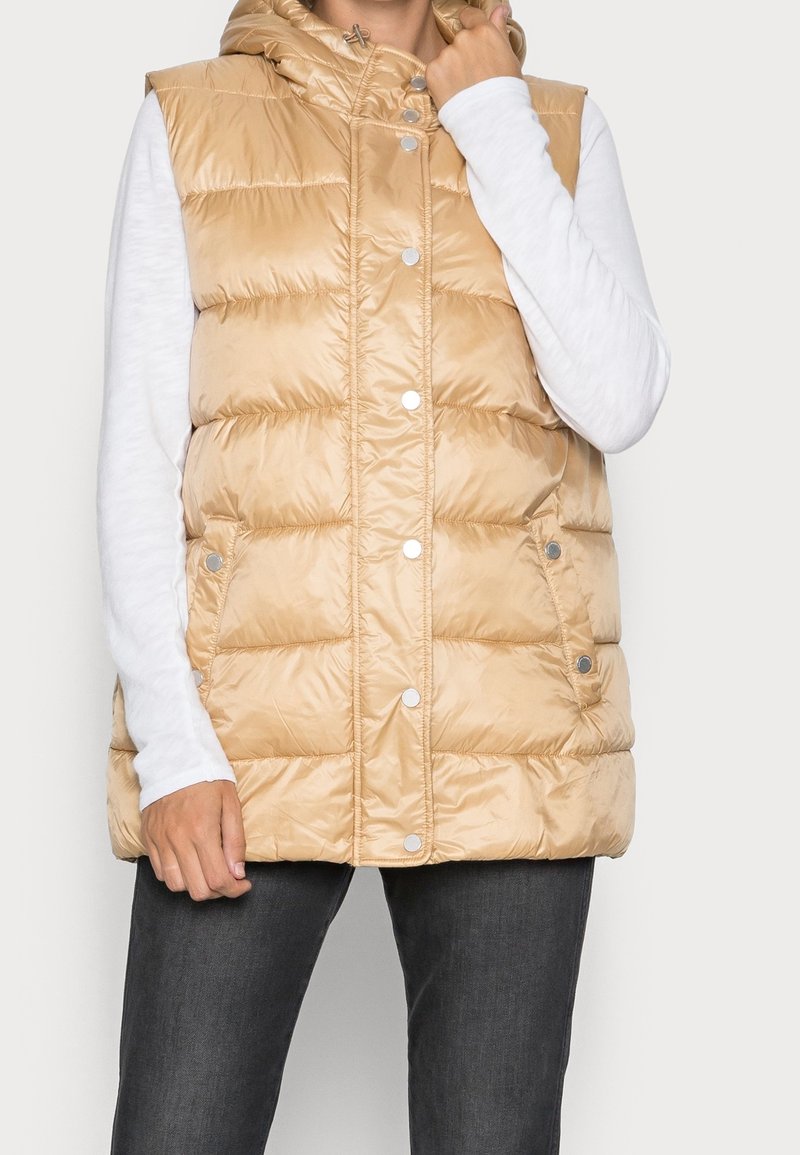 Beige pufferväst med huva, tryckknappar och sidofickor. Tillverkad av glänsande, quiltat material. Bärs över en vit långärmad skjorta.