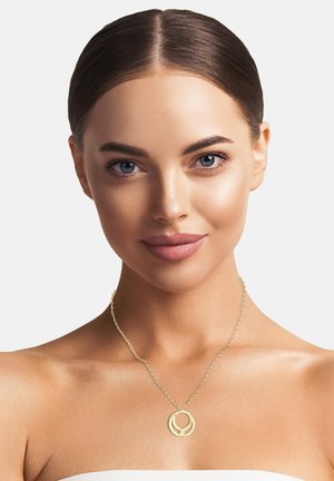 Goldene Anhänger-Halskette an einer filigranen Kette, mit einem kreisförmigen Design aus verwobenen Elementen, getragen von einem Modell mit glatter Haut.