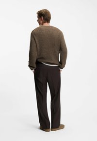 Maglione lavorato a maglia marrone con un motivo texturizzato e spesso, abbinato a pantaloni marroni scuri. L'outfit è completato da scarpe beige.