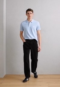 Polo shirt azzurro con colletto e piccolo logo, abbinato a pantaloni neri a gamba dritta e scarpe da ginnastica nere. Sfondo in legno chiaro e parete grigia.