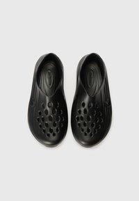 A fekete klumpák sima műanyagból készültek, slip-on kialakításúak, lekerekített formájuk van, és több szellőzőnyílás található a felső felületükön.