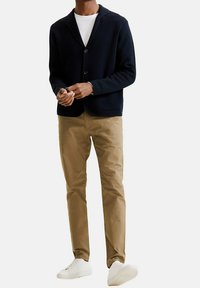 Blazer in maglina blu con colletto a scialle e doppio petto, abbinato a chinos color khaki e sneakers bianche. Il blazer ha tasche frontali.