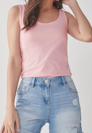 Femme portant un haut sans manches rose clair et un jean bleu clair déchiré, posant avec un bras levé près de sa tête.