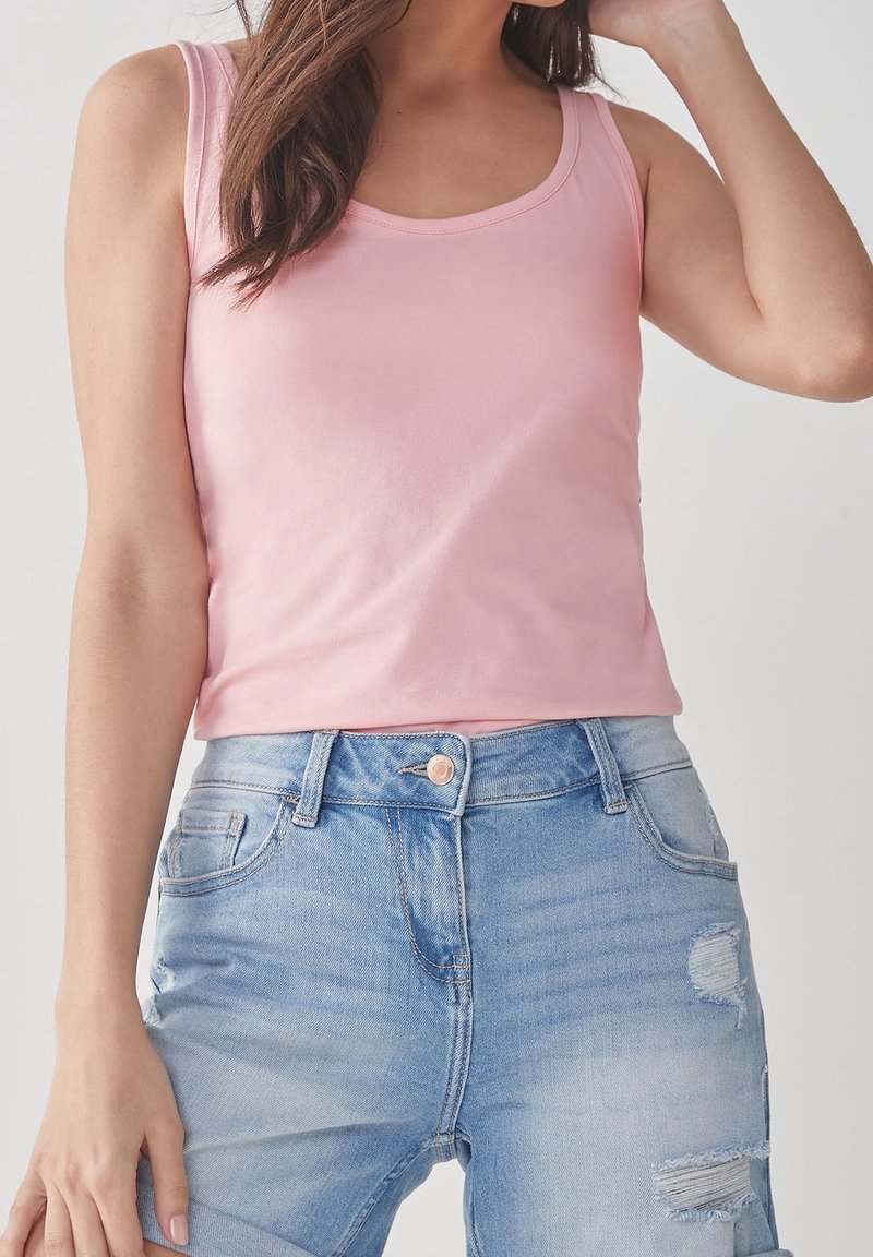 Femme portant un haut sans manches rose clair et un jean bleu clair déchiré, posant avec un bras levé près de sa tête.