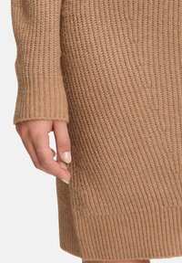 Strickpullover in einem warmen Beigeton. Verfügt über ein texturiertes Design mit gerippten Bündchen und Saum. Hand befindet sich in der Nähe des Pullovers.