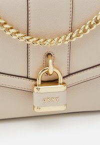 Borsa a mano in pelle beige con tracolla a catena dorata e accentuazione con lucchetto dorato. Presenta cuciture sottili e una superficie liscia. Marchio DKNY visibile.