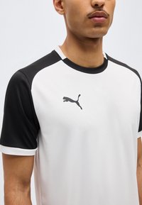 Homme portant un maillot de sport Puma blanc et noir, visible du cou au milieu du torse, sur un fond clair uni.