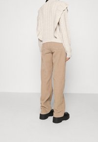 Personne portant un pantalon en velours côtelé beige, des chaussures plateformes noires, et un pull en maille torsadée crème avec un tricot assorti drapé sur les épaules, debout.