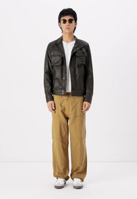 Μαύρο δερμάτινο μπουφάν με δύο μπροστινές τσέπες, λευκό μπλουζάκι, καφέ παντελόνι και γκρίζα αθλητικά. Το μοντέλο φοράει στρογγυλά γυαλιά ηλίου. Απλό σχέδιο, χαλαρή εφαρμογή.