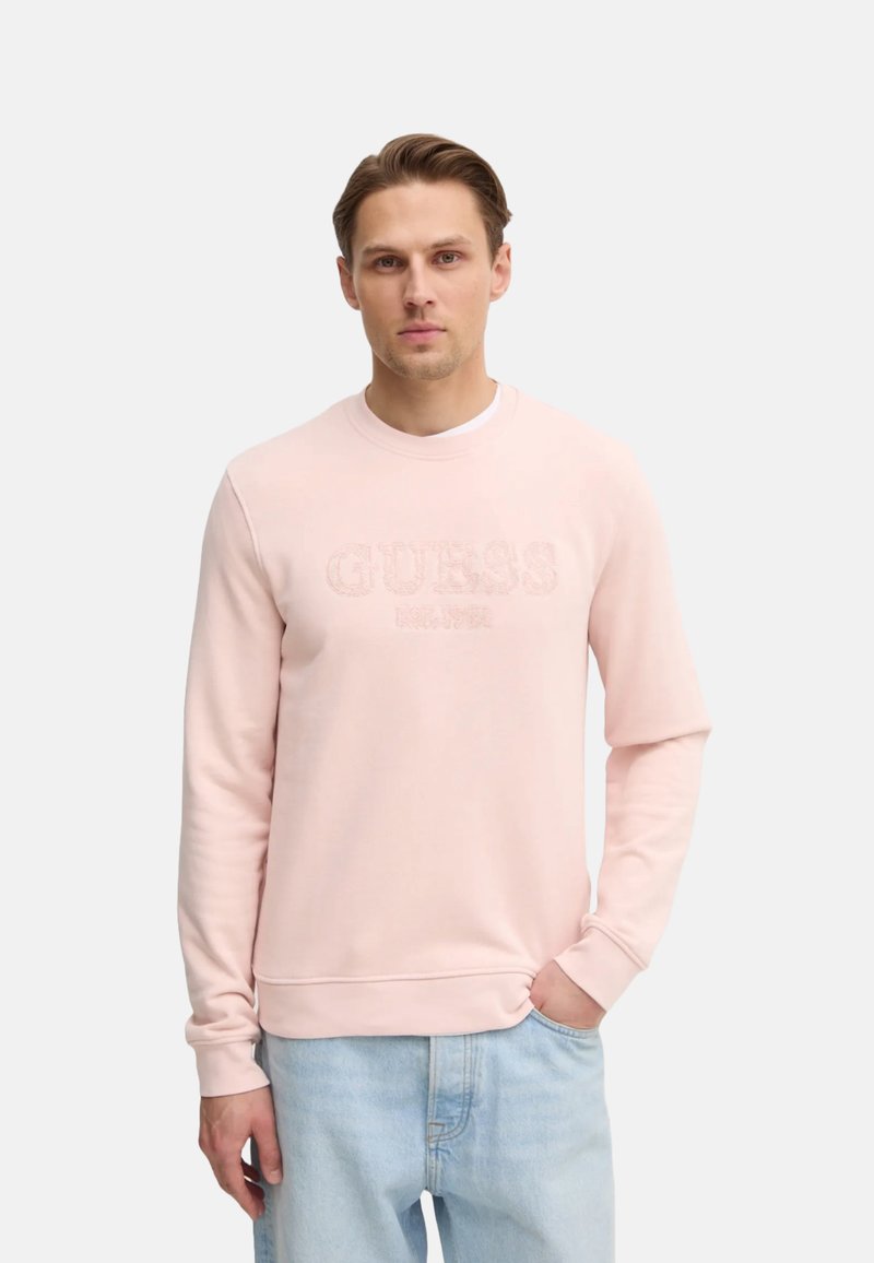 Sudadera rosa claro con cuello redondo, logo bordado "GUESS" y puños acanalados. Combinada con jeans de denim azul claro.
