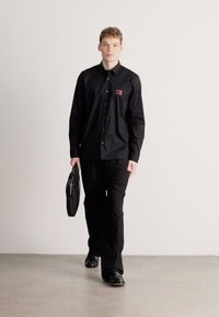 Μαύρο πουκάμισο με κουμπιά και μικρή κόκκινη ετικέτα, συνδυασμένο με μαύρο παντελόνι και παπούτσια. Το μοντέλο κρατάει μια μαύρη τσάντα και περπατάει σε ουδέτερο πάτωμα.