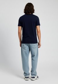 Mies, jolla on olkapäille ulottuvat hiukset, pukeutuneena tummansiniseen t-paitaan, vaaleansinisiin farkkuihin ja valkoisiin lenkkareihin, seisoo selin yksinkertaisella taustalla.