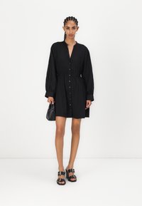 Rochie neagră, cu nasturi în față, mâneci lungi și talie elastică. Asortată cu sandale negre cu toc, ce au accente aurii, și o geantă mică neagră.