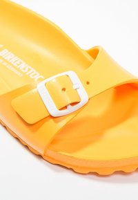 Gul Birkenstock sandal med en bred rem og en hvid spænde. Fremstillet af fleksibelt materiale med en struktureret overflade. Letvægtsdesign.