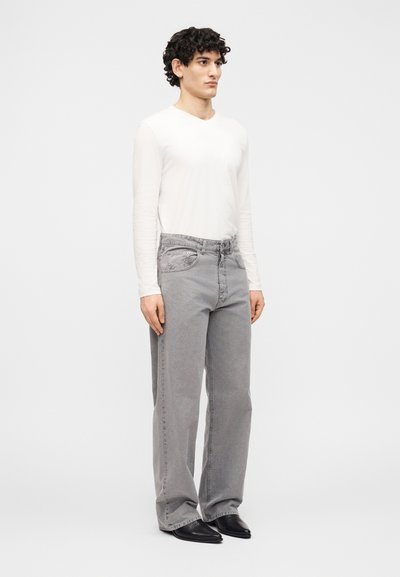 Νέος άνδρας με σγουρά μαλλιά, φορώντας λευκό μακρυμάνικο πουκάμισο, φαρδιά γκρι τζιν και μαύρα μυτερά παπούτσια, στέκεται μπροστά σε έναν απλό φόντο.