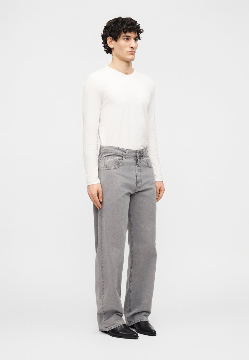 Jeune homme aux cheveux bouclés portant une chemise blanche à manches longues, un jean gris ample et des chaussures noires pointues, debout devant un fond uni.