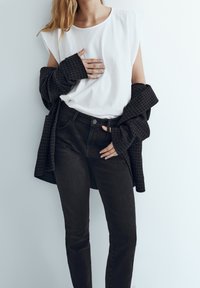 Un haut sans manches blanc associé à une chemise à carreaux sombre et un jean noir. Le tissu lisse et la coupe ajustée mettent en valeur la silhouette.