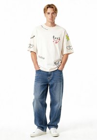 Baltas per didelis marškinėliai su įvairiais juodais ir spalvotais siuvinėjimais, derinamas su laisvomis mėlynomis džinsomis ir baltais sportbačiais.