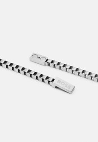 Bracelet en métal argenté avec des maillons entrelacés et un fermoir rectangulaire gravé du mot "BOSS". Design poli et élégant.