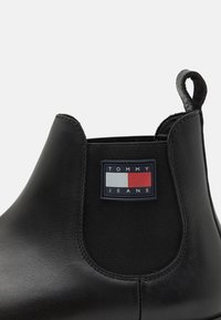 Bottines Chelsea noires en cuir lisse avec des panneaux latéraux élastiques, ornées d'une patch logo rectangulaire en marine, rouge et blanc au niveau de la cheville.