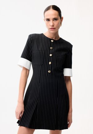 Kobieta ubrana w czarną sukienkę w prążki z białymi mankietami i złotymi guzikami, stojąca na tle jednolitego białego tła.