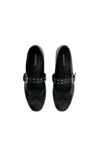 Zapatos de cuero negro con puntera redondeada, que presentan un lazo decorativo y una correa adornada con tachuelas plateadas. Textura suave, suela plana.