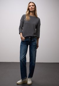 Femme debout, les mains dans les poches, portant un pull à rayures noires et blanches, un jean bleu et des baskets blanches, sur un fond uni.