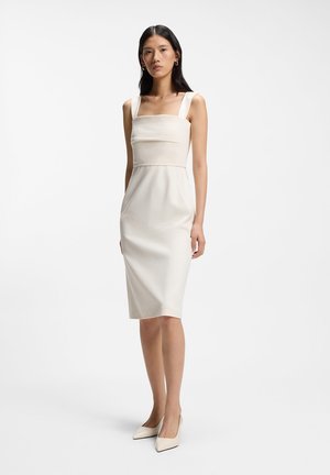 Woman standing wearing a sleeveless, knee-length, fitted cream dress and matching pointed-toe heels against a plain white background.