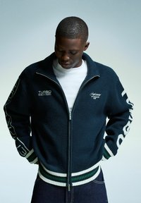 Jeune homme regardant vers le bas, portant un pull zippé bleu marine avec des côtes rayées blanches et vertes et un texte blanc sur la poitrine et les manches.