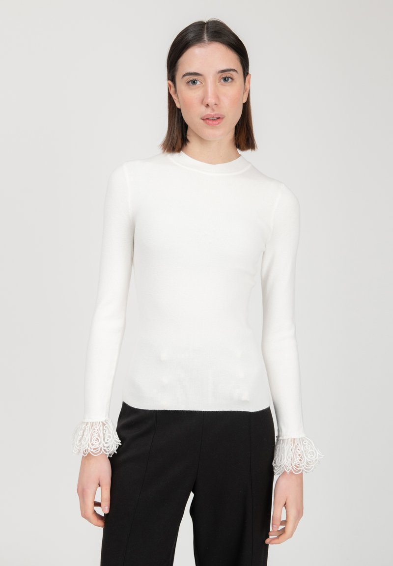 Maglietta bianca a maniche lunghe con un alto colletto, caratterizzata da polsini in pizzo trasparente con motivi intricati, abbinata a pantaloni neri.