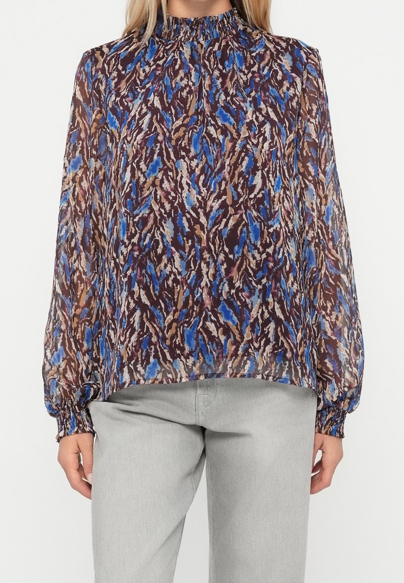 Femme portant un chemisier transparent à manches longues avec un motif abstrait bleu et marron, col haut froncé, associé à un pantalon gris clair.