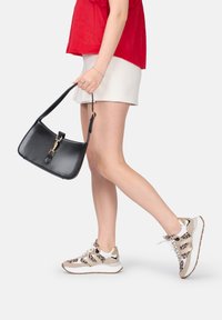 Borsa a tracolla nera in pelle con hardware dorato, tenuta con una mano. Il modello indossa un top rosso, una gonna bianca e sneakers con stampa leopardata.