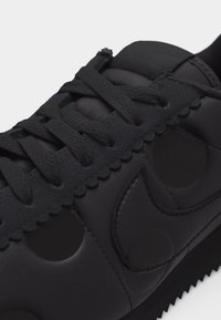 Μαύρο αθλητικό παπούτσι με κυματιστές άκρες, στρογγυλές κοπές, υφασμένα κορδόνια και μεγάλο λογότυπο Swoosh στο πλάι.