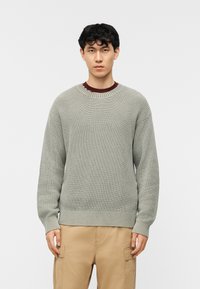 Νέος άνδρας στέκεται μπροστά σε απλό φόντο φορώντας ανοιχτό γκρι πλεκτό πουλόβερ πάνω από μπορντό πουκάμισο και μπεζ παντελόνι cargo.