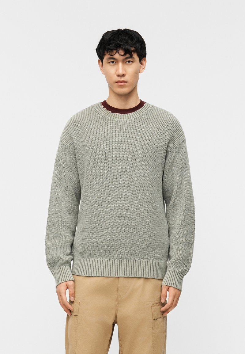 Νέος άνδρας στέκεται μπροστά σε απλό φόντο φορώντας ανοιχτό γκρι πλεκτό πουλόβερ πάνω από μπορντό πουκάμισο και μπεζ παντελόνι cargo.