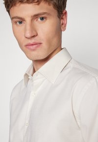 Camicia bianca a trama con colletto strutturato, caratterizzata da sottili motivi geometrici e bottoni simili a perle, indossata da un modello.