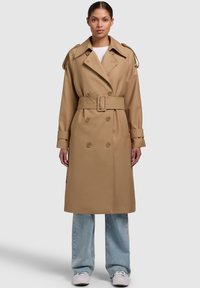 Beige trench coat s dvouvrstvým designem, pásem v pase, širokými klopami a ramenními epauletami. Nosí se přes světle modré džíny.