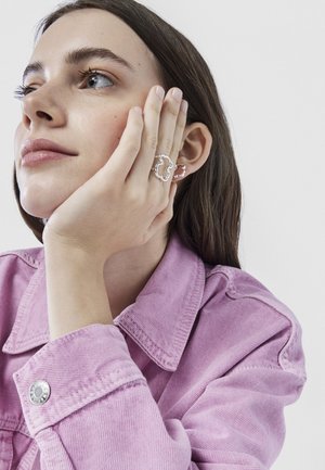 Anillo de metal plateado con una forma abstracta de nube, llevado en la mano, acompañado de un pequeño pendiente. La modelo lleva una chaqueta de mezclilla rosa.